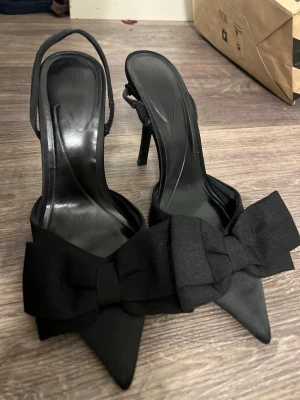 Svarta pumps med stor rosett - Säljer ett par eleganta svarta pumps med spetsig tå och hög klack. Skorna har en stor dekorativ rosett framtill och öppen häl med smal rem. Materialet ser ut att vara satin eller liknande tyg som ger en lyxig känsla.