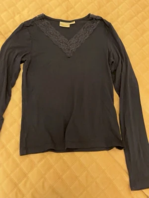 Mörkblå långärmad topp med spets - Mörkblå långärmad topp med v-ringning dekorerad med spetsdetalj. Tunn och mjuk i materialet, perfekt för lager på lager eller som den är. Enkel men ändå elegant design som passar till mycket.