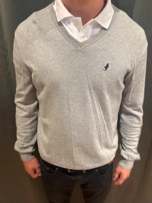 Marlboro classic V-Neck - Snygg ljusgrå V-ringad tröja med diskret broderad logga på bröstet. Tröjan har långa ärmar och ribbade muddar vid ärmslut och nederkant. Perfekt att bära över en skjorta för en clean och stilren look.