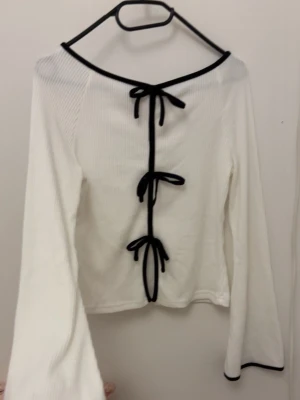 Vit långärmad topp med svarta knyt - Trendig vit långärmad topp med svarta kanter och tre dekorativa knyt framtill. Toppen har en lätt ribbad struktur och vida ärmar, vilket ger en cool och unik look. Perfekt för dig som vill sticka ut lite extra.