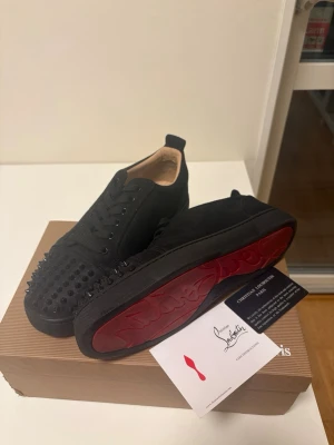 Svarta sneakers med nitar och röd sula - Säljer ett par svarta sneakers från Christian Louboutin med coola svarta nitar på tån och den ikoniska röda sulan. Skorna har snörning och är tillverkade i mocka med läderdetaljer. Perfekta för dig som vill sticka ut med din stil.