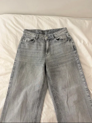 Ljusgrå raka jeans med hög midja - Säljer ett par ljusgrå jeans med raka ben och hög midja. Klassisk femficksmodell med bälteshällor och knappgylf. Jeansen har en avslappnad passform och är perfekta för en trendig vardagslook.