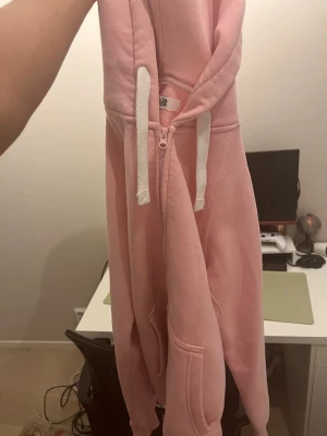 Rosa hoodie - Säljer en ljusrosa hoodie med dragkedja och vita snören i huvan. Hoodien har fickor framtill och är tillverkad i ett mjukt material som känns skönt mot huden. Perfekt för en chill och avslappnad stil.