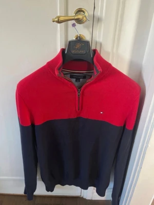 Röd och marinblå tröja Tommy Hilfiger - Snygg tvåfärgad tröja från Tommy Hilfiger med röd ovandel och marinblå nederdel. Tröjan har halv dragkedja, ribbad krage och diskret logga på bröstet. Perfekt för dig som gillar klassisk stil med en modern twist.