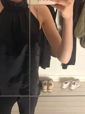 Svart halterneck linnetopp - Säljer en svart halterneck linnetopp i ett glansigt material. Toppen är ärmlös och har en hög halsringning som ger en clean och stilren look. Perfekt att styla med både jeans och kjol för en trendig vibe.