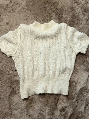 Vit stickad topp med kort ärm - Säljer en söt vit stickad topp med korta ärmar och hög ribbad midja. Tröjan har ett diskret mönster i stickningen och en mjuk, ribbad halsringning. Perfekt att styla med jeans eller kjol för en trendig look.