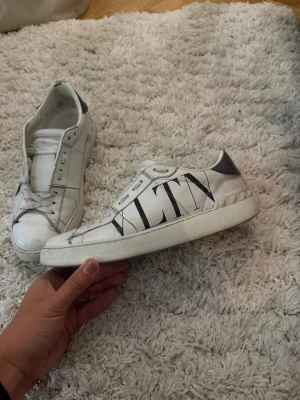Vita Valentino VLTN sneakers - Säljer ett par vita sneakers från Valentino med stor VLTN-logga på sidan. Skorna har svarta detaljer vid hälen och är tillverkade i läder. Klassisk låg modell med chunky sula och coola nitar baktill. Perfekta för dig som gillar stilrena men ändå statement skor.