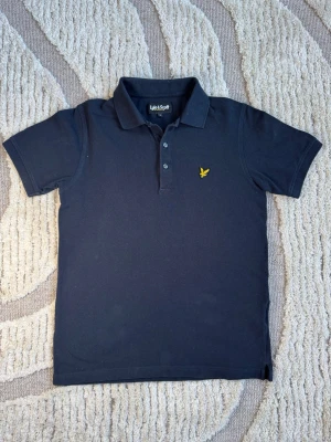 Marinblå piké från Lyle & Scott - Snygg marinblå pikétröja från Lyle & Scott med klassisk krage, korta ärmar och knappslå framtill. Bröstet pryds av den ikoniska gula loggan. Tillverkad i bomull för en skön känsla och stilren look.