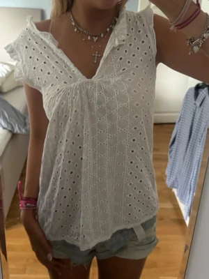 Vit super snygg blus ❣️ - Söt vit blus med broderade hålmönster och volangdetaljer vid axlarna. Blusen är ärmlös och har en djup V-ringning framtill. Tillverkad i ett lätt och luftigt tyg, perfekt för sommardagar. Passar till både jeans och kjol för en trendig look.🥰