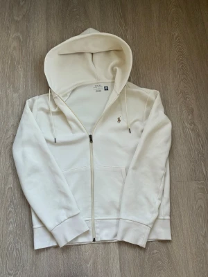 Vit hoodie med dragkedja från Polo Ralph Lauren - Snygg vit hoodie från Polo Ralph Lauren med dragkedja framtill och klassisk huva med dragsko. Broderad logga i beige på bröstet. Tillverkad i mjukt bomullsmaterial och har ribbade muddar vid ärmslut och nederkant. Perfekt för en clean och stilren look.