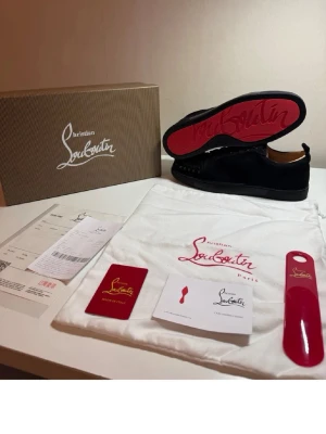 Svarta Christian Louboutin sneakers - Säljer ett par svarta sneakers från Christian Louboutin med klassisk röd sula och svarta nitar på ovandelen. Skorna har en låg profil och är tillverkade i mocka med svarta skosnören. Perfekt för dig som vill sticka ut med exklusiv design.
