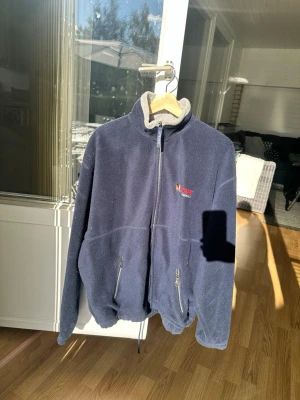 Marinblå fleecejacka med dragkedja - Mysig marinblå fleecejacka med hög krage och hel dragkedja framtill. Två praktiska fickor med dragkedja och broderad logga på bröstet. Perfekt för kyliga dagar och enkel att slänga på sig över en hoodie.