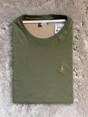 Olivgrön t-shirt från Polo Ralph Lauren - Snygg olivgrön t-shirt från Polo Ralph Lauren med klassisk rund hals och broderad logga på bröstet. T-shirten är i mjuk bomull och har en enkel, stilren design som passar perfekt till jeans eller shorts. Storlek s men är lite liten i storleken 