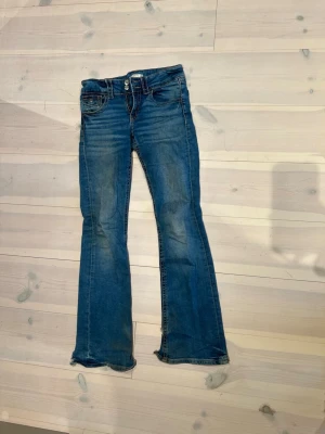 Blå bootcut jeans från Gina Tricot - Snygga blå bootcut jeans från Gina Tricot med dubbla knappar och klassiska fickor. Jeansen har en lätt tvättad look och utsvängda ben, perfekta för en trendig och avslappnad stil. Tvättas innan de skickas.
