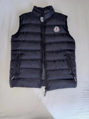 Svart dunväst från Moncler - Snygg svart dunväst från Moncler med quiltad design och hög krage. Västen har två dragkedjeförsedda fickor och Moncler-logga på bröstet. Perfekt för lager-på-lager och håller dig varm med stil. Helt oanvänd och bra kvalité 