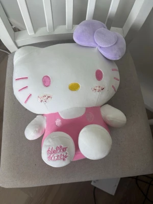 Stor Hello Kitty mjukisdjur - OBS ! Glittret är påsatt själv, försvinner med 99% säkerhet i tvätt. Ca 30x30cm gulligt Hello Kitty mjukisdjur med vit kropp, rosa overall och broderad text på foten. Den har ett stort lila rosett på huvudet och söta rosa detaljer i ansiktet. Perfekt som dekoration eller kramkompis för alla Hello Kitty-fans!