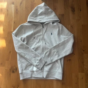  Polo Ralph Lauren hoodie  - Snygg ljusgrå hoodie med dragkedja från Polo Ralph Lauren. Klassisk design med huva och dragsko, samt den ikoniska lilla loggan broderad på bröstet. Tillverkad i mjukt bomullsmaterial som känns skönt mot huden. Perfekt för en avslappnad och stilren look.   