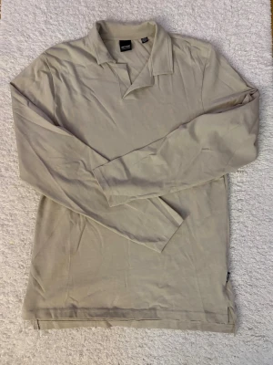 Beige långärmad pikétröja - Stilren beige långärmad pikétröja med klassisk krage och enkel design. Tillverkad i mjukt bomullstyg som känns skönt mot huden. Perfekt för dig som gillar en clean och avslappnad look.