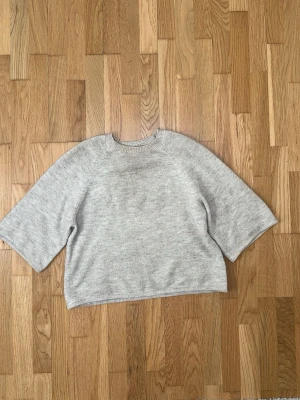 Beige stickad tröja med vida ärmar - Mysig ljusgrå stickad tröja med rund halsringning och trekvartsvida ärmar. Tröjan har en avslappnad passform och är tillverkad i ett mjukt material som passar perfekt för kyligare dagar.