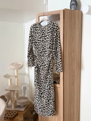 Lång leopardmönstrad klänning - Säljer en långärmad klänning med leopardmönster i svart och beige. Klänningen har rund halsringning och rak passform. Öppen i ryggen och liten slits baktill. 