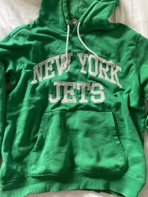Grön New York Jets hoodie - Snygg grön hoodie med stor vit text 'NEW YORK JETS' på bröstet. Klassisk känguruficka framtill och justerbar huva med vita snören. Perfekt för dig som gillar sportig stil och vill sticka ut lite extra. Den är använd men väl omhändertagen.