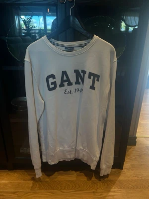 Vit sweatshirt från GANT XL - Klassisk vit sweatshirt från GANT med svart tryck 'GANT Est. 1949' på bröstet. Tröjan har rund halsringning, långa ärmar och ribbade muddar vid ärmslut och nederkant. Perfekt för en avslappnad och stilren look. Går att pruta!