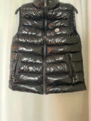 Svart glansig dunväst från Moncler - Snygg svart dunväst från Moncler med glansig finish och klassisk logotyp på bröstet. Västen har två dragkedjeförsedda fickor och hög krage. Perfekt för lager-på-lager och ger en trendig look till din outfit.