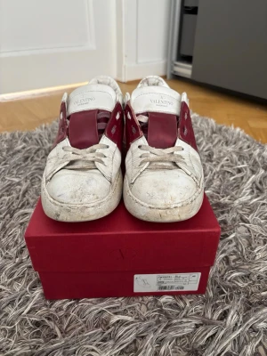 Valentino vita och röda sneakers - Säljer ett par vita sneakers från Valentino med röda detaljer på sidorna och tungan. Skorna har klassisk låg profil, vita snören och är tillverkade i läder. Kommer med originalkartong och extra skosnören.
