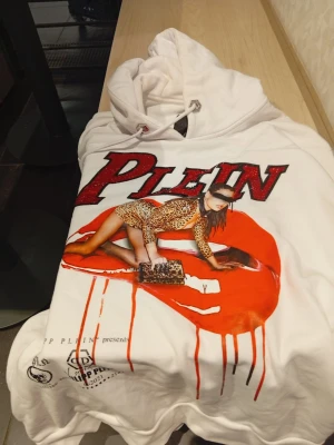 Vit Philipp Plein hoodie med tryck(Limited Edition från 2022) - Säljer en vit hoodie från Philipp Plein med stor röd och svart logga samt ett färgstarkt tryck av en modell i leopardmönstrad outfit. Hoodien har huva med dragsko och coola detaljer framtill. Perfekt för dig som gillar statement-plagg och streetwear vibes. Mycket bra skick!