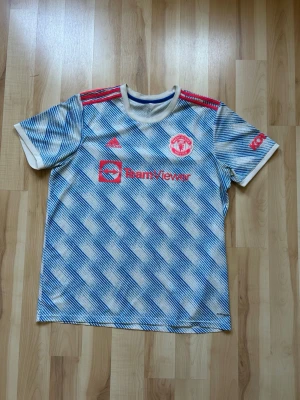 Manchester United bortatröja blå/vit - Säljer en Manchester United bortatröja från Adidas i blått och vitt med diagonalrandigt mönster. Tröjan har röda detaljer, klubbmärke och sponsorlogga på bröstet samt röda axelränder. Tillverkad i lätt och ventilerande material, perfekt för fotboll eller träning.