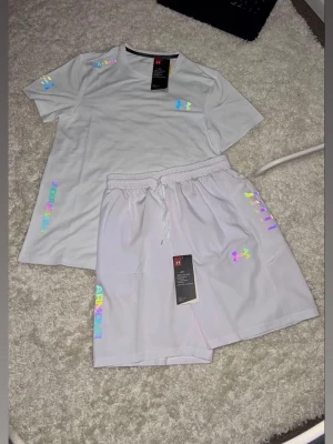 Vit träningsset från Under Armour - Säljer ett fräscht vitt träningsset från Under Armour med t-shirt och shorts. Setet har holografiska loggor och detaljer, samt elastisk midja med dragsko på shortsen. Perfekt för gymmet eller löprundan.