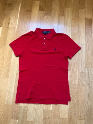 Polo Ralph Lauren piké  - Klassisk röd pikétröja från Polo Ralph Lauren med blå broderad logga på bröstet. Tröjan är i bra skick,  7/10 Storlek - 14/16 years   Priset kan diskutera! Tveka inte vid frågor eller funderingar!