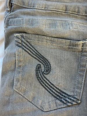 Ljusgrå jeans från Gina Tricot - Säljer ett par ljusgrå jeans från Gina Tricot med snygg broderad detalj på bakfickan. Klassisk femficksmodell med raka ben och silverfärgad knapp. Tillverkade i mjukt denimtyg som är skönt att bära hela dagen.