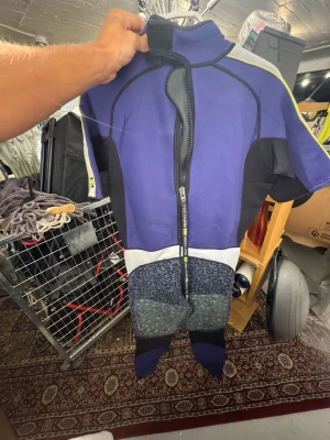 Lila våtdräkt från Helly Hansen - Kortärmad våtdräkt i lila, svart och grått från Helly Hansen. Dräkten har dragkedja framtill, hög krage med kardborrestängning och detaljer i gult. Tillverkad i tjockt neoprenmaterial som håller värmen vid vattensport. Snygg logga på bröstet och benet. Finns att hämta i Gröndal. 