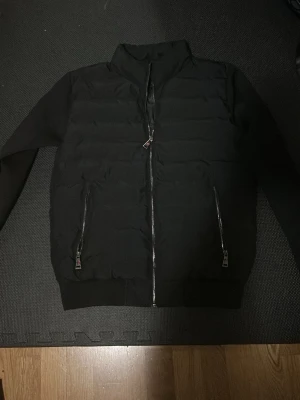 Moncler Cardingan - Säljer en svart cardigan med quiltad front och två snedställda fickor med dragkedjor. Jackan har hög krage och ribbade muddar vid ärmslut och nederkant. Materialet är en mix av vadderad polyester och mjukare tyg på ärmarna. Perfekt för kalla vinterdagar.