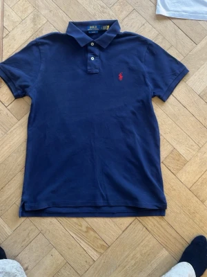 Marinblå pikétröja från Ralph Lauren - Klassisk marinblå pikétröja från Ralph Lauren med två knappar och röd broderad logga på bröstet. Tröjan har korta ärmar och mjukt kragmaterial, perfekt för en avslappnad men stilren look.
