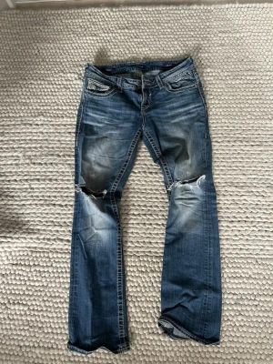 Blå slitna bootcut jeans - Säljer ett par blå bootcut jeans med slitningar och hål på knäna. Jeansen har ljusa tvättade partier och kontrastsömmar. Klassisk femficksmodell och låg midja. Perfekta för en avslappnad och trendig look. Köptes för 700kr. 