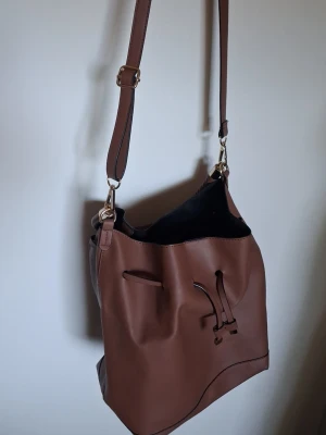 Brun bucket bag med axelrem - Snygg brun bucket bag i slätt konstläder med justerbar axelrem och flätat handtag. Väskan har dragskostängning och minimalistisk design, perfekt för dig som gillar stilrena accessoarer. Rymmer allt du behöver till vardagen.