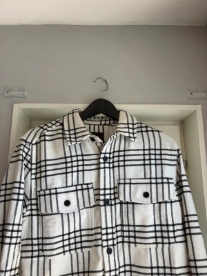 Vit och svart rutig overshirt H&M - Säljer en snygg vit overshirt från H&M med svart rutmönster. Skjortjackan har två stora bröstfickor med knappar, klassisk krage och knäppning framtill. Perfekt lager på lager-plagg i mjukt tyg som ger en avslappnad look.