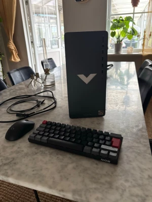 HP Victus stationär dator med tillbehör - Säljer en HP Victus stationär dator i svart med stilren design och vitt V-logotyp på fronten. Medföljer kompakt mekaniskt tangentbord med svarta och röda tangenter samt en svart gamingmus. Perfekt för gaming eller studier. Flera USB-portar och moderna anslutningar på baksidan. Hör gärna av er om ni har frågor👍🏻
