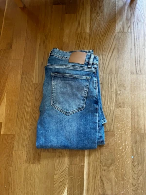 Blå raka jeans från GANT - Säljer ett par klassiska blå jeans från GANT med rak passform och femficksmodell. Jeansen är endast använda fåtal gånger. Tillverkade i slitstark denim med läderpatch bak i midjan.innerbenslängd : 84cm. Midja 44cm. Hör av dig vid funderingar! 