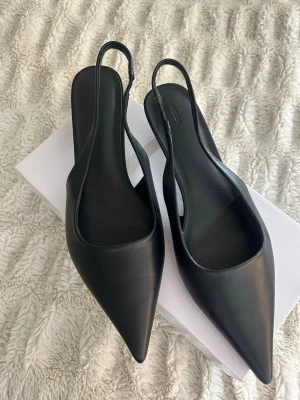 Svarta slingbacks med låg klack - Svarta klackskor med spetsig tå och slingback-rem, aldrig använda, från Nelly i storlek 39. Skorna har låg klack och är tillverkade i slätt material som ger en elegant look. 