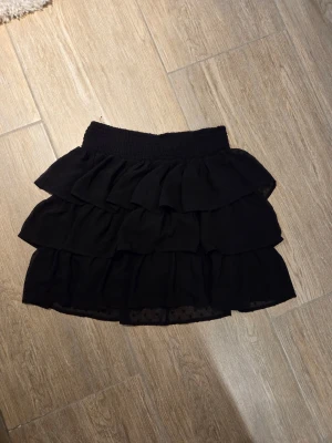 Svart volangkjol med resårmidja - Kort svart kjol med flera lager volanger och bred resår i midjan. Kjolen har en luftig och flowy känsla, perfekt för att styla med en enkel topp eller stickad tröja. Materialet känns lätt och bekvämt.