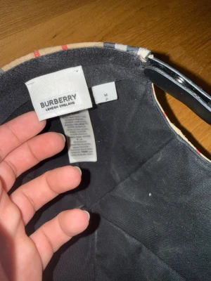 Burberry keps med klassiskt rutmönster - Snygg keps från Burberry med det ikoniska beige, svarta och röda rutmönstret. Justerbar bak med svart plastspänne och tydlig Burberry London England-logga. Tillverkad i bomull för en bekväm känsla och stilren look.