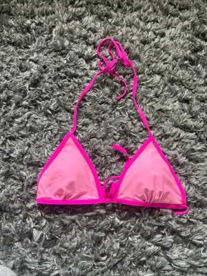 Rosa trekantsbikini med knyt - Snygg trekantsbikini i stark rosa färg med knyt i nacken och ryggen. Bikinin har tunna band och klassisk triangelmodell, perfekt för sommaren. Materialet känns som syntet och ger en skön passform vid stranden eller poolen.