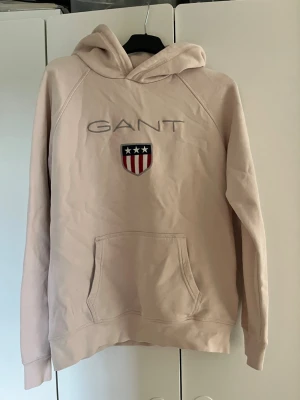 Rosa hoodie från GANT med logga - Snygg rosa hoodie från GANT med broderad logga och sköld med stjärnor och ränder på bröstet. Hoodien har huva, känguruficka och ribbade muddar. Perfekt för en avslappnad och trendig look. Storlek 158/164