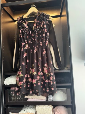 Svart blommig kortklänning med volang - Supersöt svart kortklänning med rosa blommönster och volangdetaljer vid axlar och urringning. Klänningen har v-ringad hals och smock i midjan för en bekväm passform. Perfekt för vår och sommar!
