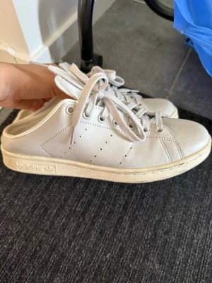 Vita Adidas Stan Smith sneakers - Klassiska vita Adidas Stan Smith sneakers i läder med perforerade ränder på sidorna och vit sula. Skorna har snörning och diskret logga på sidan och plösen. Perfekta för en clean och stilren look.