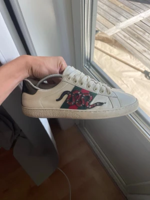 Vita sneakers med orm och ränder - Snygga vita sneakers i läder med gröna och röda sidoremsor samt en broderad orm på sidan. Skorna har vita snören och en låg profil. Perfekta för dig som vill sticka ut med coola detaljer och klassisk design.