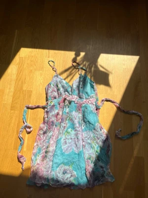 Blommig klänning med knytband - Söt kortklänning med blommigt mönster i turkos, lila och rosa toner. Klänningen har smala axelband, v-ringning och knytband i midjan storlek M 